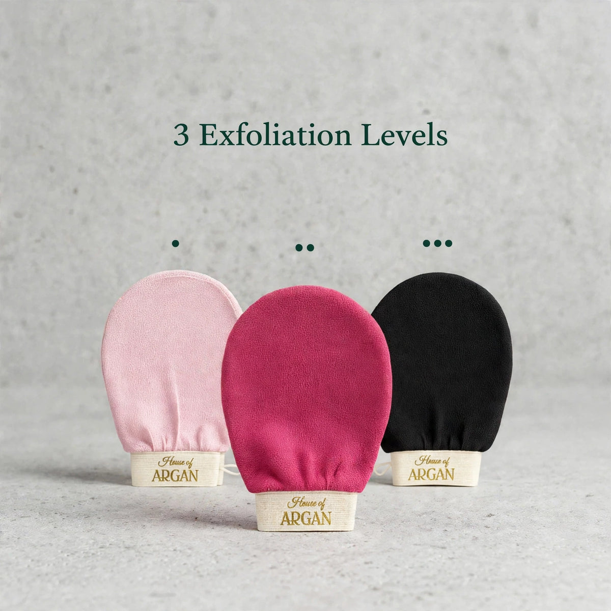 Three House of Argan kessa exfoliation gloves in pink, fuchsia, and black, displayed on a stone surface representing gentle, medium, and deep exfoliation levels